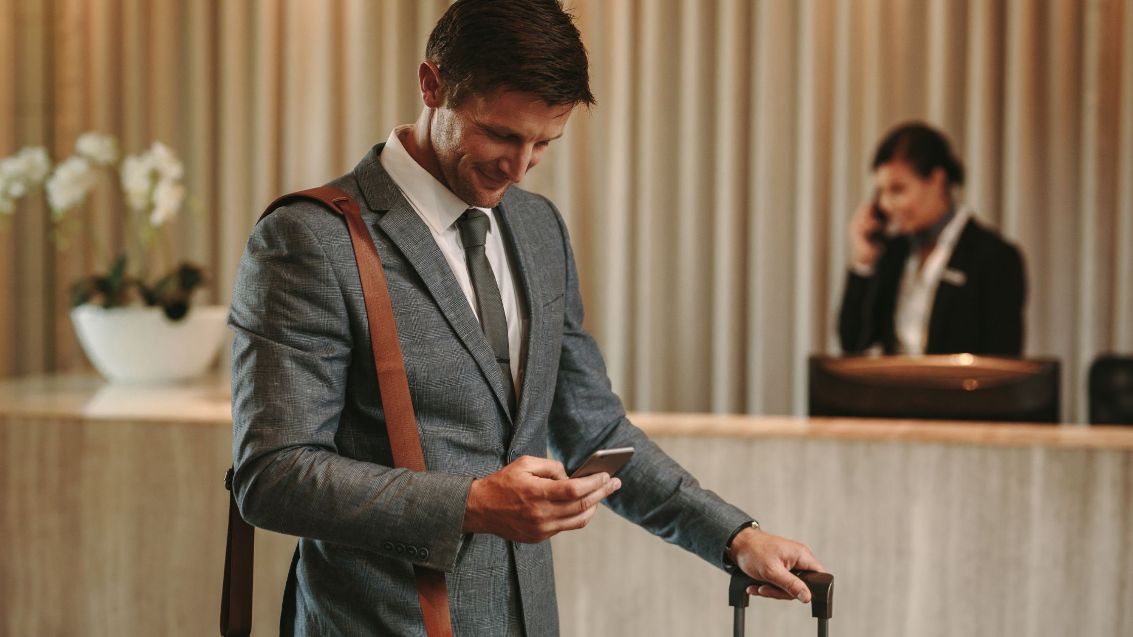 a business traveler in grey suit holding a mobile phone on one hand and suitcase handle on another in a hotel lobby, Hari's Court